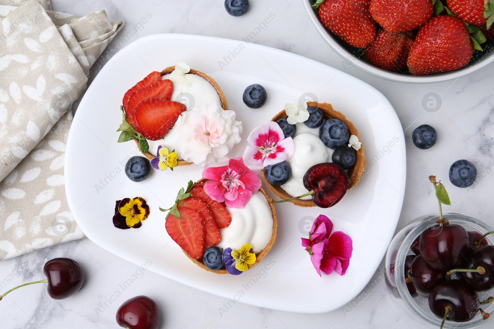 Photo of Sweet tartlets with berries and flowers on light marble table, flat lay. Delicious dessert