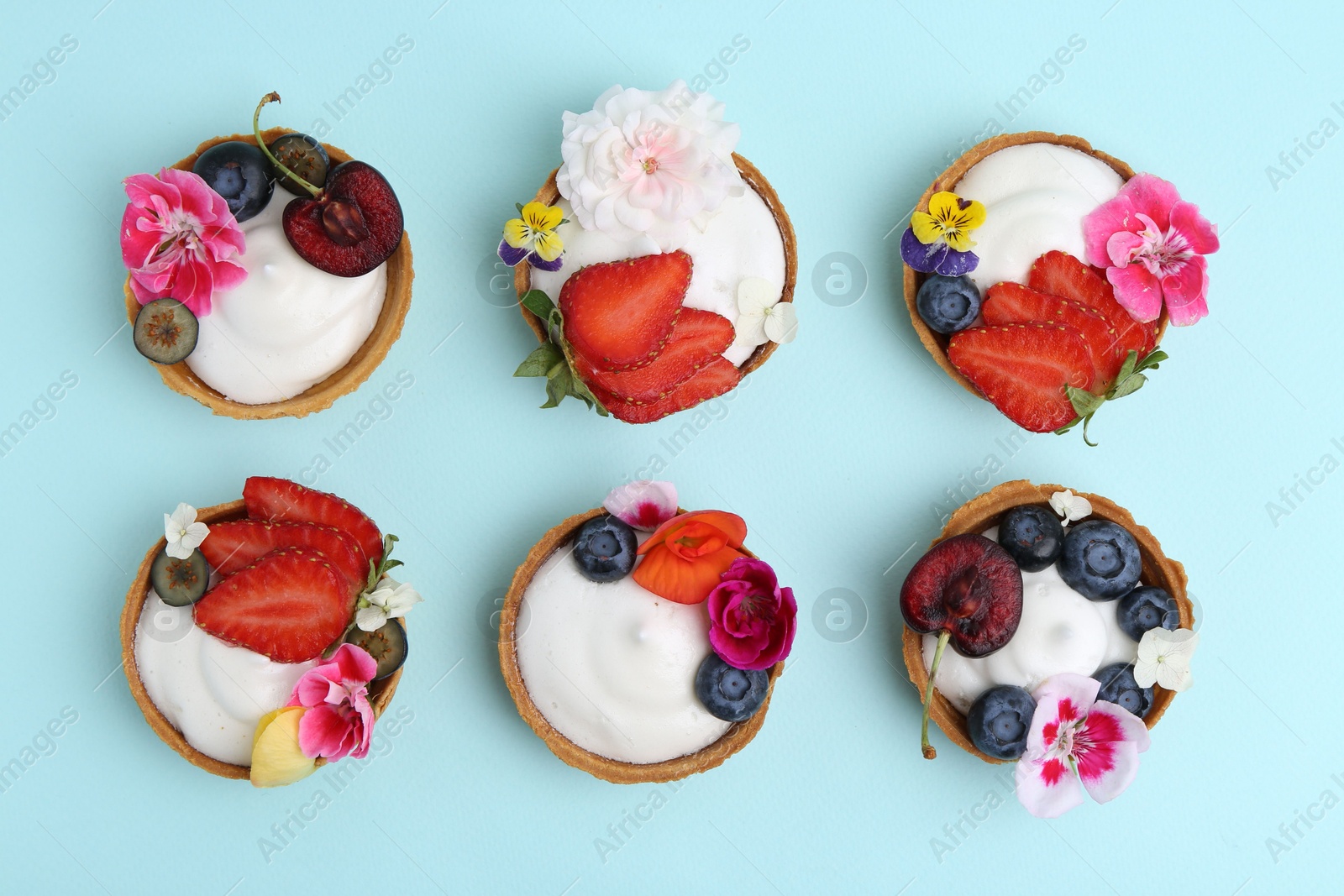 Sweet tartlets with berries and flowers on turquoise background, flat lay. Delicious dessert Photo of Sweet tartlets with berries and flowers on turquoise background, flat lay. Delicious dessert