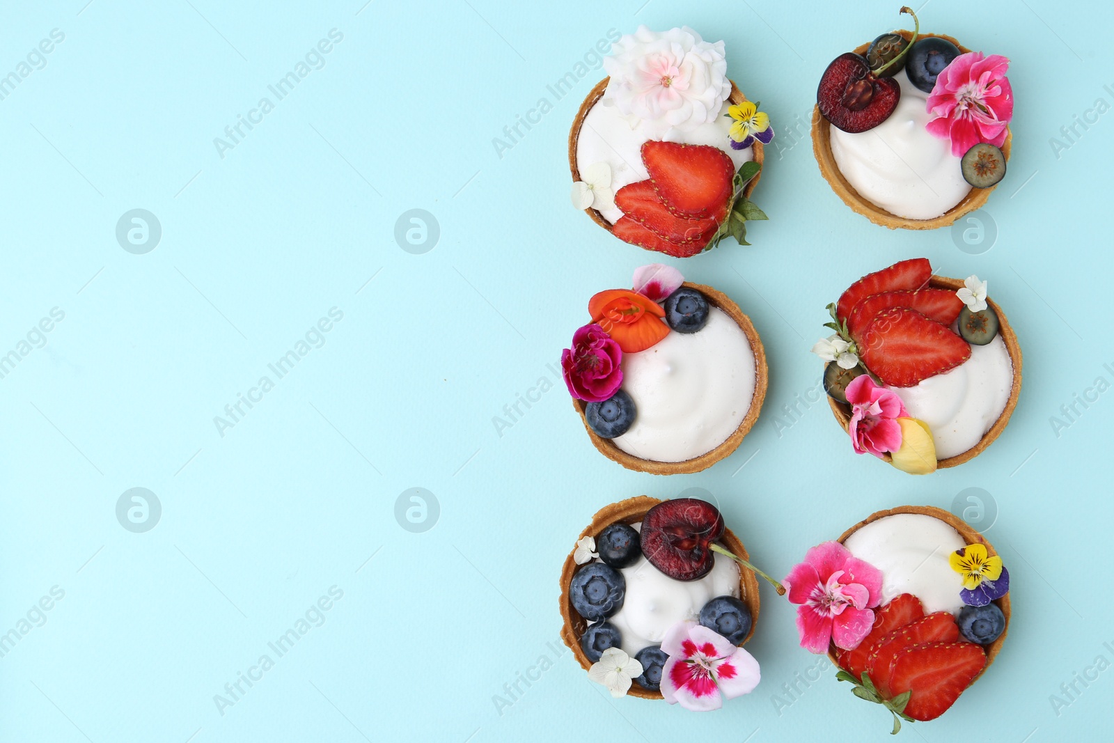 Sweet tartlets with berries and flowers on turquoise background, flat lay with space for text. Delicious dessert Photo of Sweet tartlets with berries and flowers on turquoise background, flat lay with space for text. Delicious dessert
