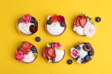 Photo of Sweet tartlets with berries and flowers on yellow background, flat lay. Delicious dessert
