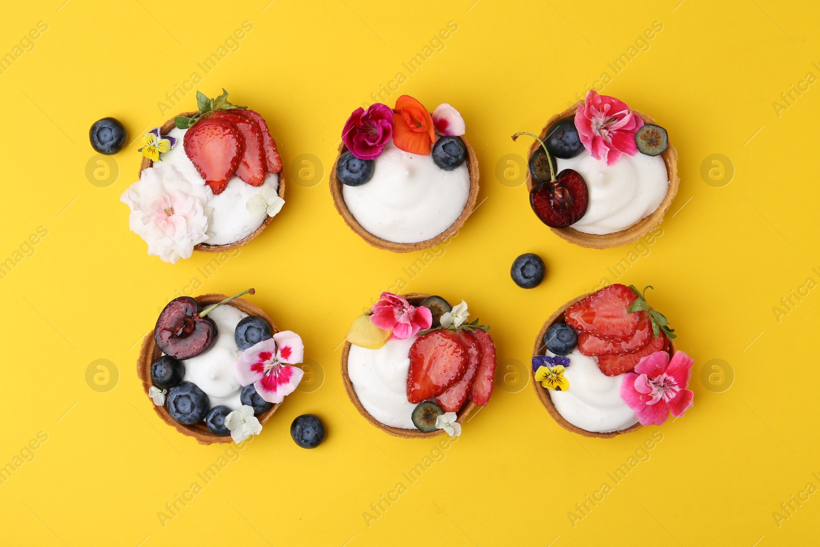 Photo of Sweet tartlets with berries and flowers on yellow background, flat lay. Delicious dessert