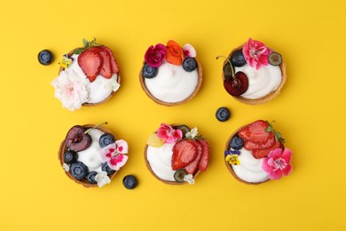 Photo of Sweet tartlets with berries and flowers on yellow background, flat lay. Delicious dessert
