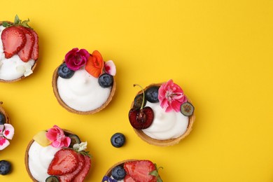Photo of Sweet tartlets with berries and flowers on yellow background, flat lay with space for text. Delicious dessert