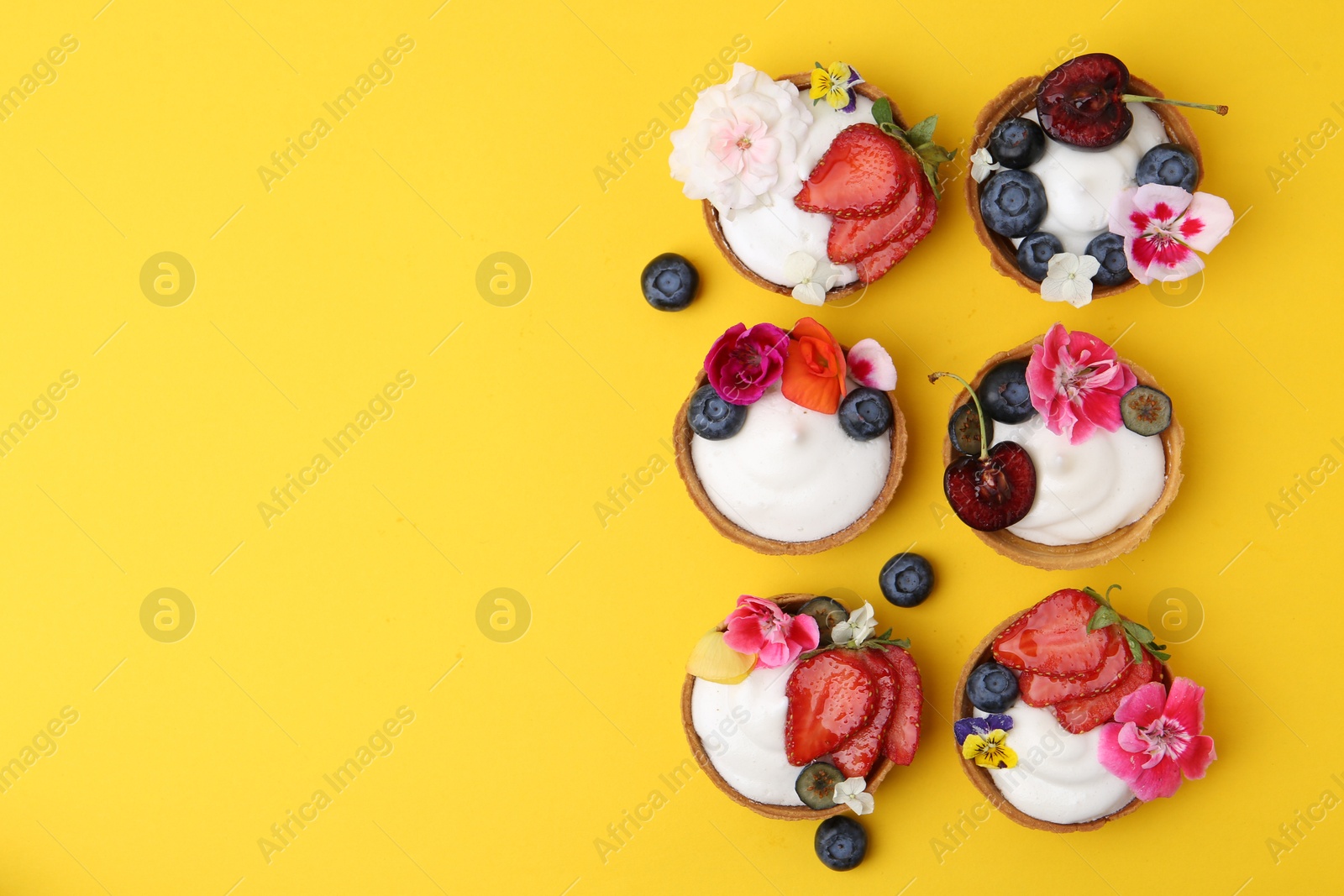Photo of Sweet tartlets with berries and flowers on yellow background, flat lay with space for text. Delicious dessert