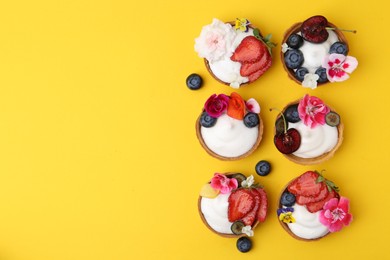 Photo of Sweet tartlets with berries and flowers on yellow background, flat lay with space for text. Delicious dessert