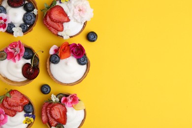 Photo of Sweet tartlets with berries and flowers on yellow background, flat lay with space for text. Delicious dessert
