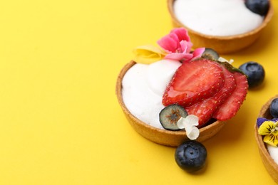 Photo of Sweet tartlets with berries and flowers on yellow background, closeup with space for text. Delicious dessert