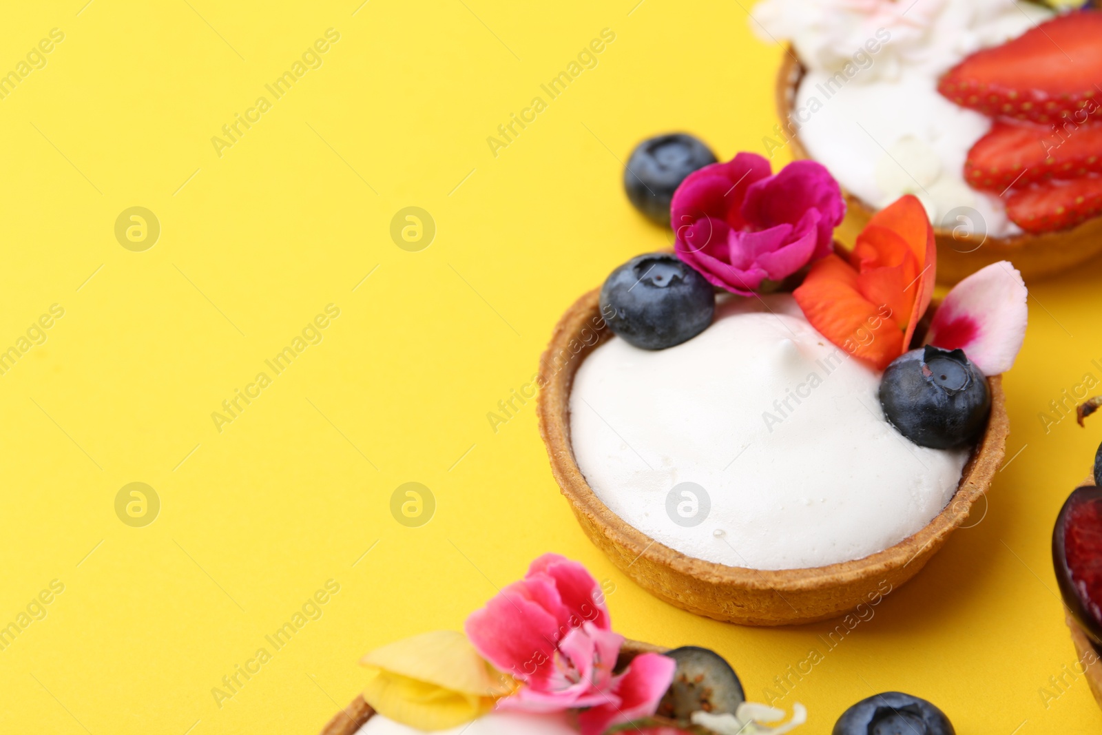 Photo of Sweet tartlets with berries and flowers on yellow background, closeup with space for text. Delicious dessert