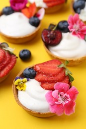 Photo of Sweet tartlets with berries and flowers on yellow background, closeup. Delicious dessert