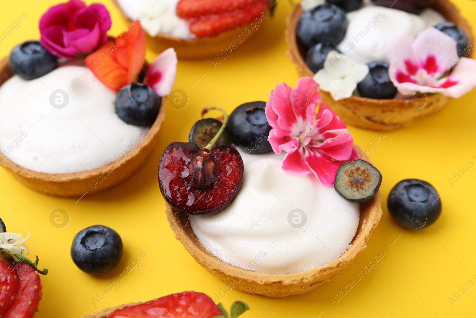 Sweet tartlets with berries and flowers on yellow background, closeup. Delicious dessert Photo of Sweet tartlets with berries and flowers on yellow background, closeup. Delicious dessert