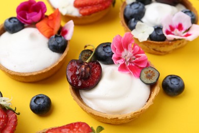 Photo of Sweet tartlets with berries and flowers on yellow background, closeup. Delicious dessert