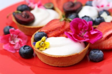Photo of Sweet tartlets with berries and flowers on red plate, closeup. Delicious dessert