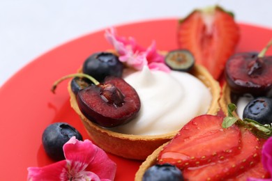 Photo of Sweet tartlets with berries and flowers on red plate against light background, closeup. Delicious dessert