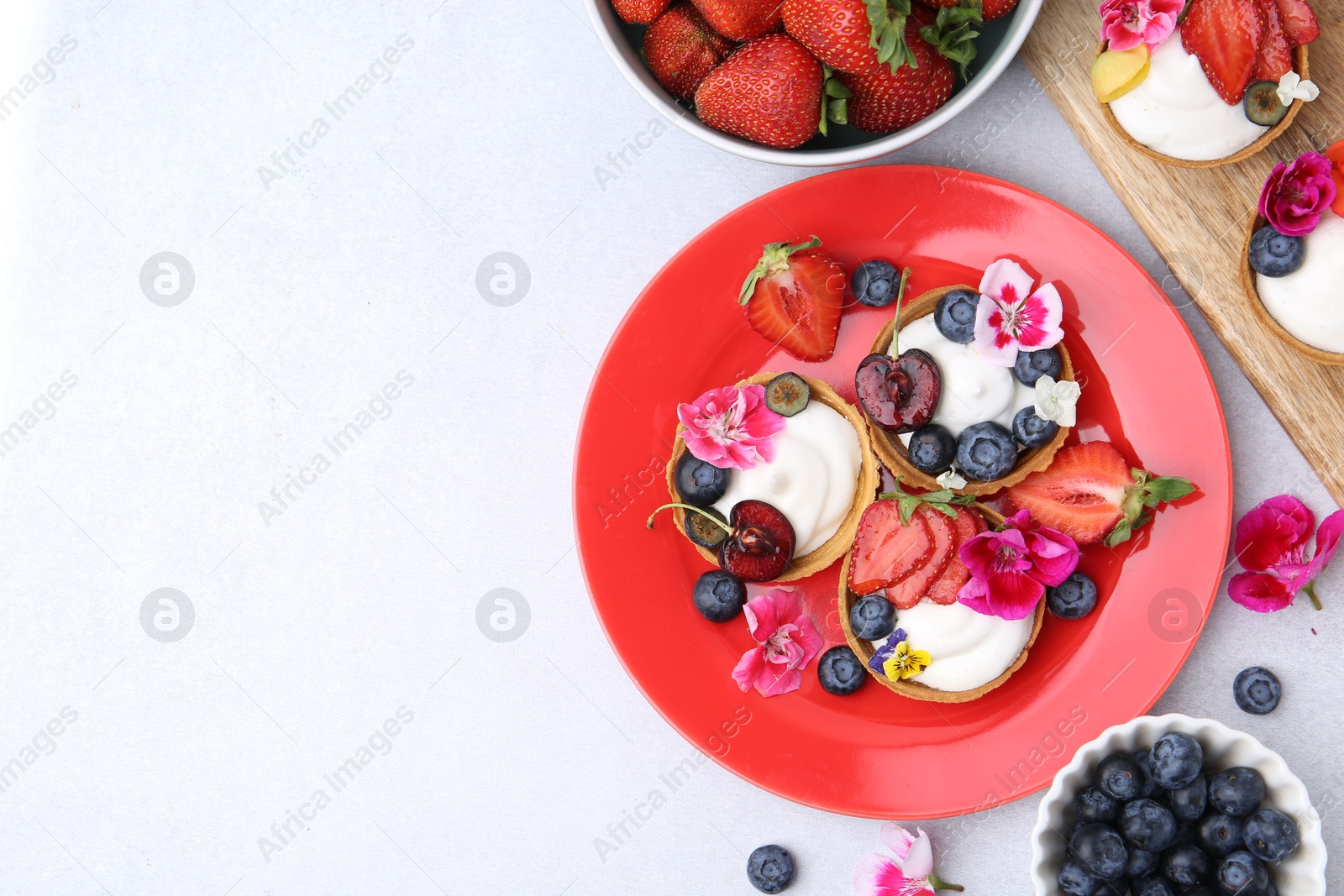 Photo of Sweet tartlets with berries and flowers on light grey table, flat lay with space for text. Delicious dessert