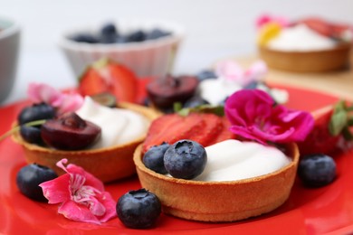 Photo of Sweet tartlets with berries and flowers on plate against light background, closeup. Delicious dessert