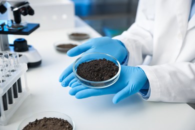 Photo of Scientist holding Petri dish with soil sample at white table indoors, closeup