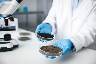 Photo of Scientist holding Petri dishes with soil samples at white table indoors, closeup