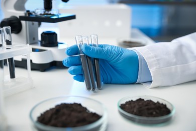 Photo of Scientist with test tubes of soil samples at white table in laboratory, closeup
