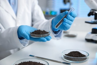Photo of Scientist holding Petri dish and test tube with soil samples at white table indoors, closeup