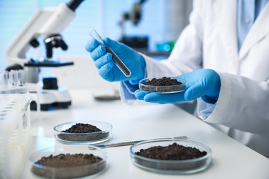 Photo of Scientist holding Petri dish and test tube with soil samples at white table indoors, closeup