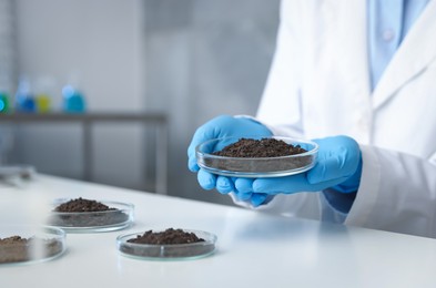 Photo of Scientist holding Petri dish with soil sample at white table indoors, closeup