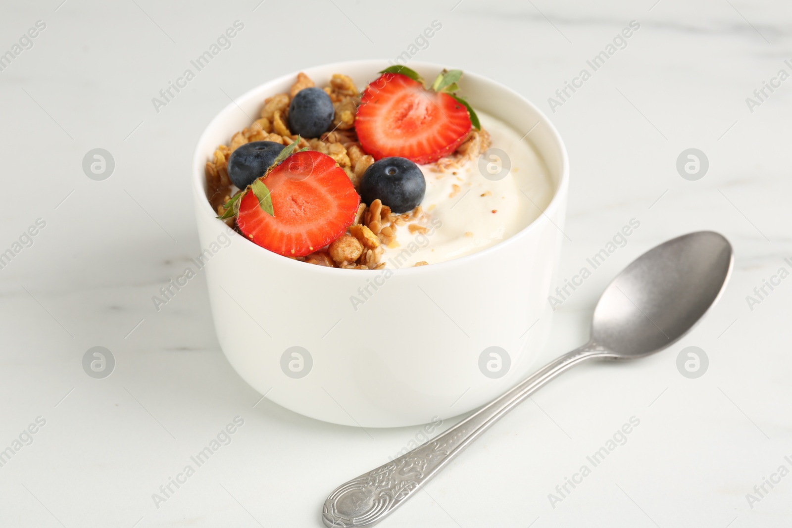 Photo of Tasty yogurt with granola, berries and spoon on white marble table, closeup