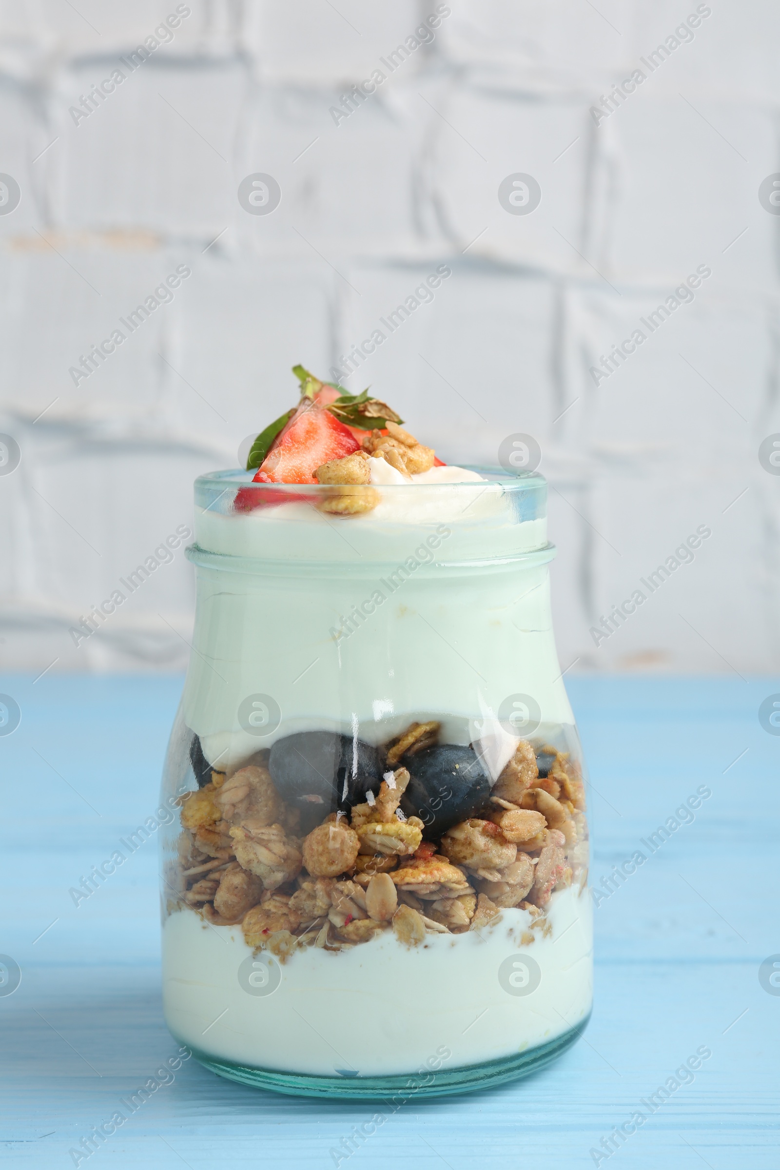 Photo of Tasty yogurt with granola and berries in glass jar on light blue wooden table, closeup