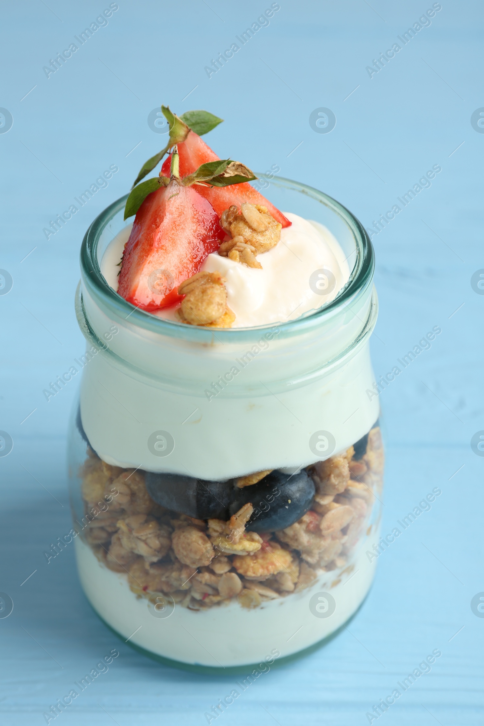 Photo of Tasty yogurt with granola and berries in glass jar on light blue wooden table, closeup