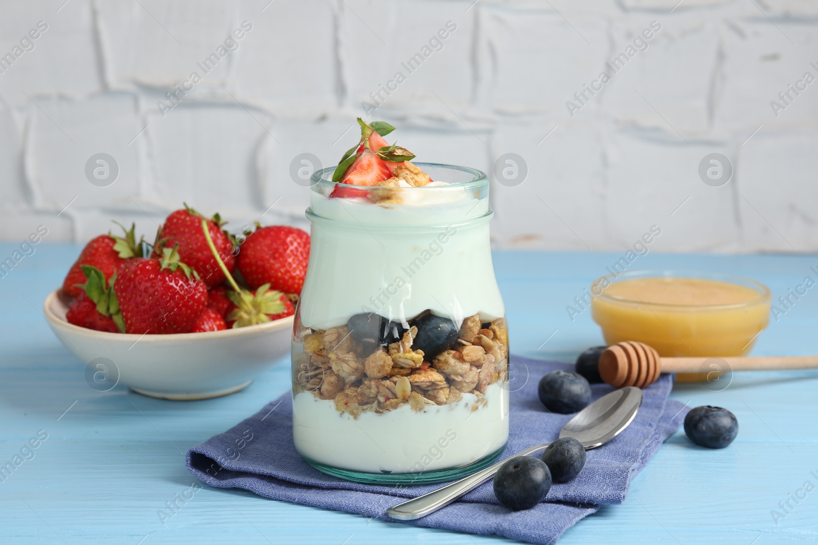 Tasty yogurt with granola, berries, honey and spoon on light blue wooden table Photo of Tasty yogurt with granola, berries, honey and spoon on light blue wooden table