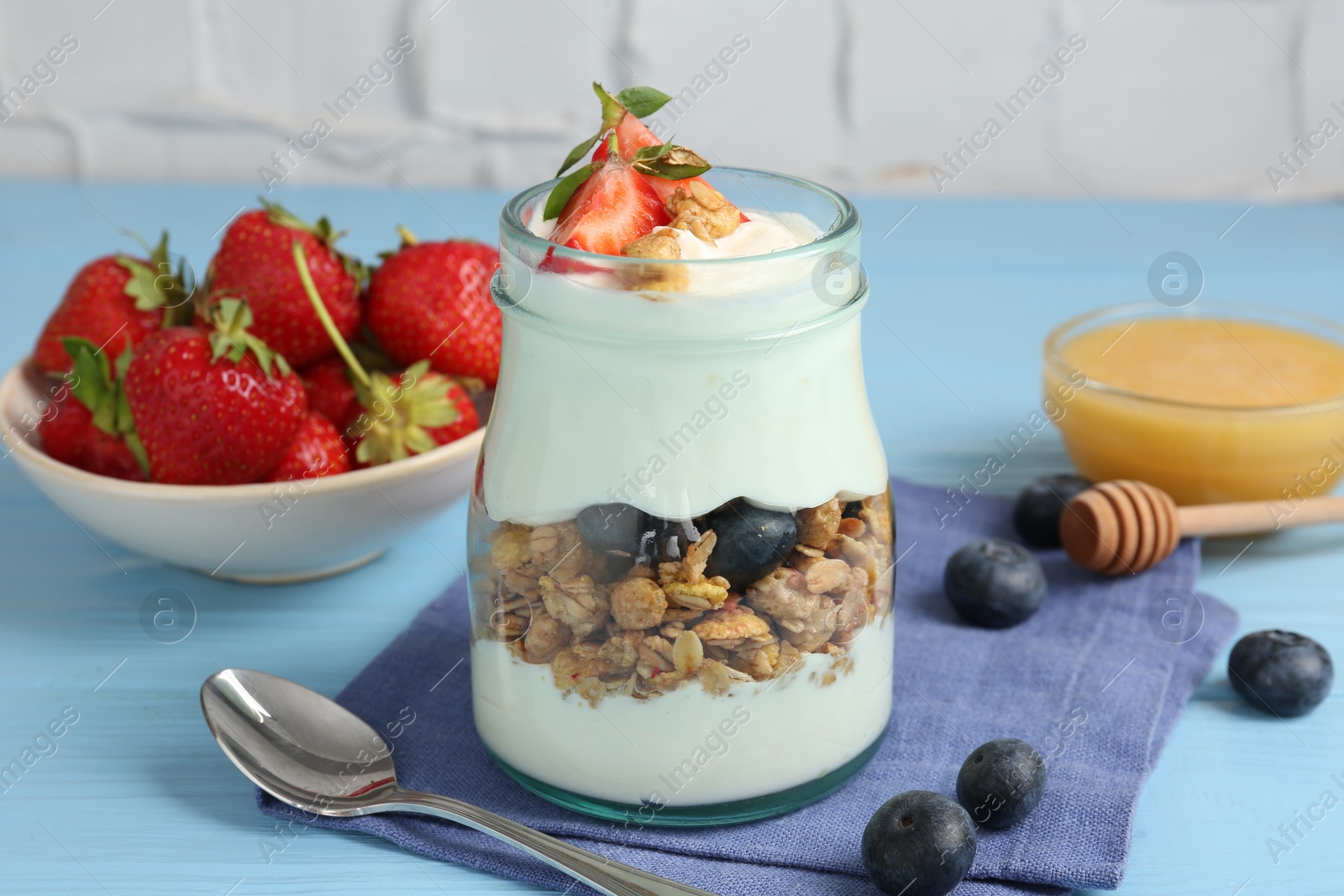 Photo of Tasty yogurt with granola, berries, honey and spoon on light blue wooden table, closeup