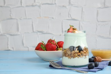 Photo of Tasty yogurt with granola, berries, honey and spoon on light blue wooden table, space for text