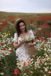 Photo of Smiling woman in hat with bag of chamomile flowers walking in wildflower meadow