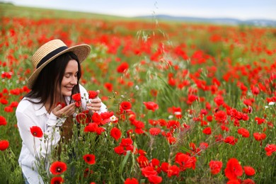 Photo of Smiling woman in hat sniffing red poppy flowers in wildflower meadow