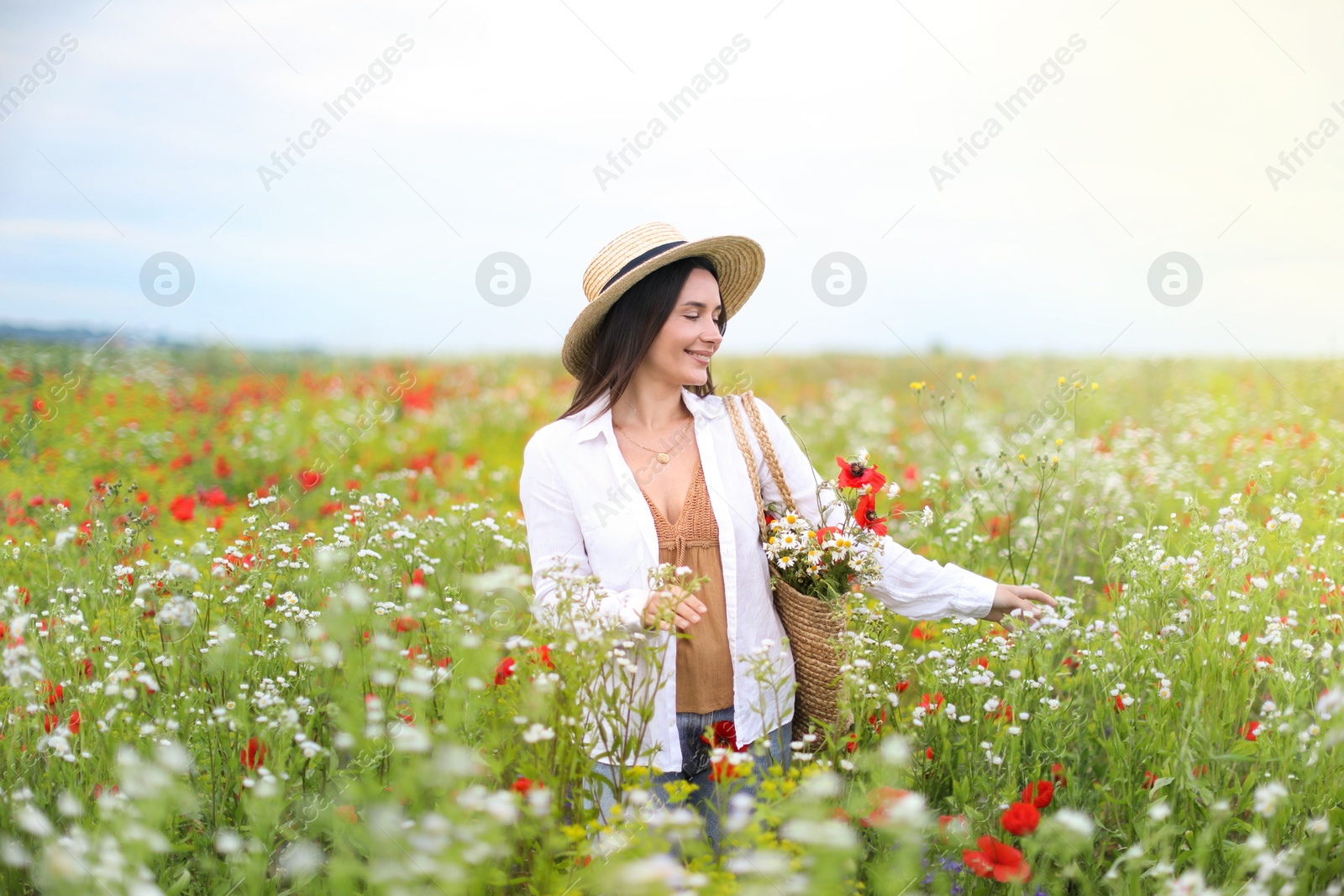 Photo of Smiling woman in hat with bag of flowers walking in wildflower meadow
