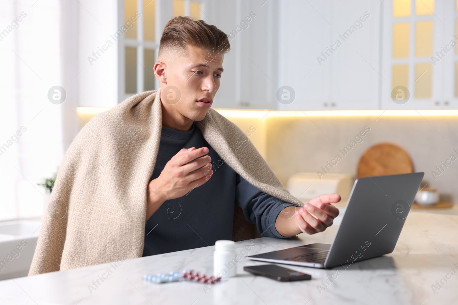 Photo of Sick man having online consultation with doctor via laptop at white marble table in kitchen