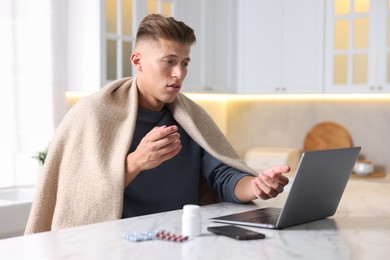 Photo of Sick man having online consultation with doctor via laptop at white marble table in kitchen