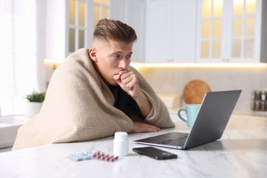 Photo of Sick man having online consultation with doctor via laptop at white marble table in kitchen