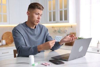 Photo of Sick man with pills having online consultation with doctor via laptop at white marble table in kitchen