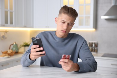 Photo of Sick man with pills having online consultation with doctor via smartphone at white marble table in kitchen