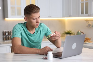 Photo of Sick man with thermometer having online consultation with doctor via laptop at white marble table in kitchen