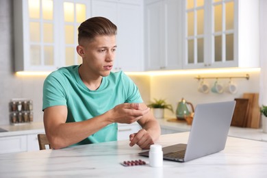 Photo of Sick man having online consultation with doctor via laptop at white marble table in kitchen