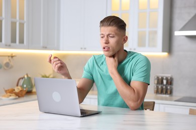 Photo of Sick man having online consultation with doctor via laptop at white marble table in kitchen