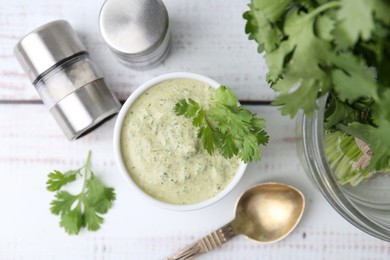 Photo of Tasty cilantro sauce in bowl, spoon and shakers on white wooden table, flat lay