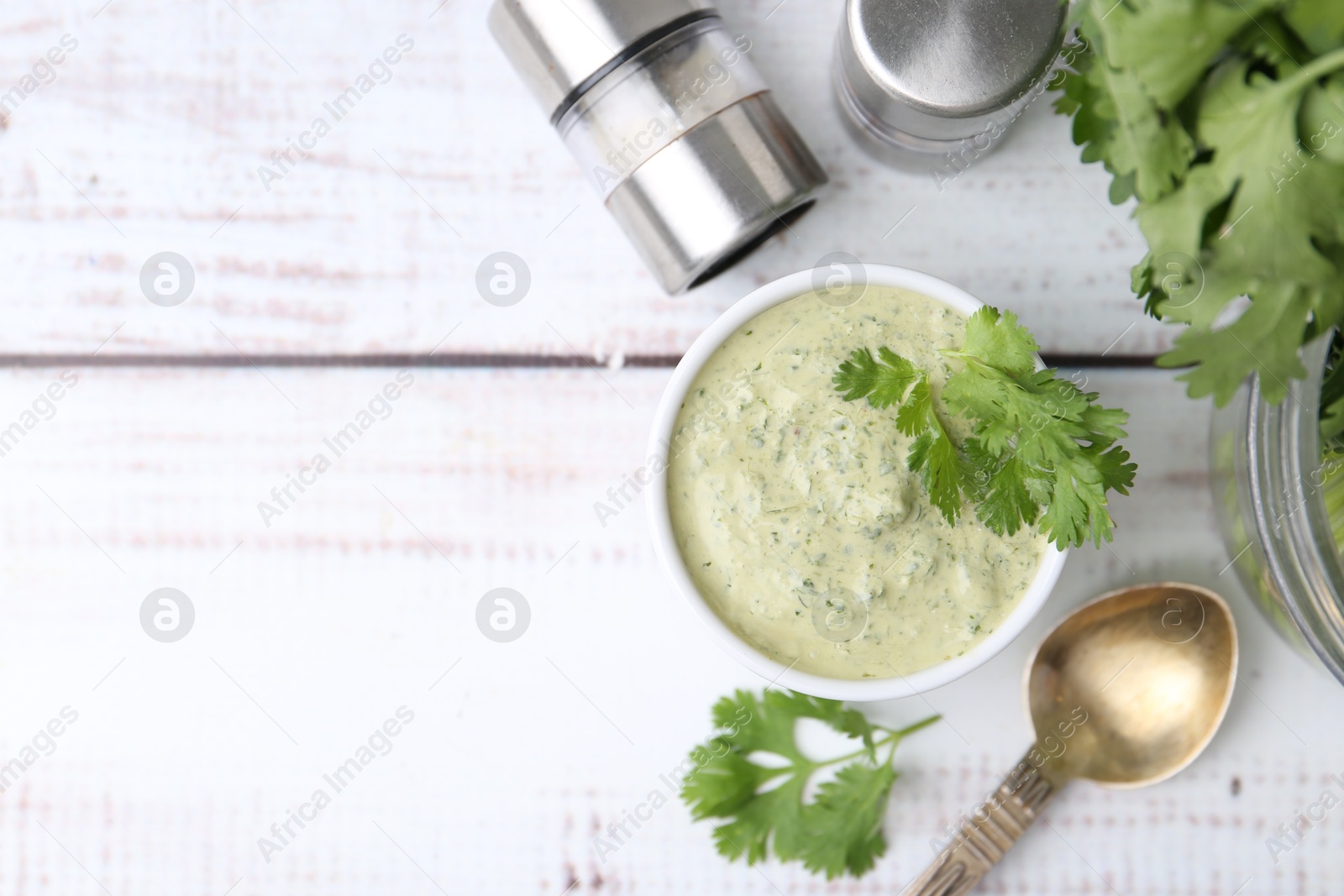 Photo of Tasty cilantro sauce in bowl, spoon and shakers on white wooden table, flat lay. Space for text