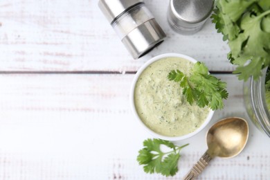 Photo of Tasty cilantro sauce in bowl, spoon and shakers on white wooden table, flat lay. Space for text