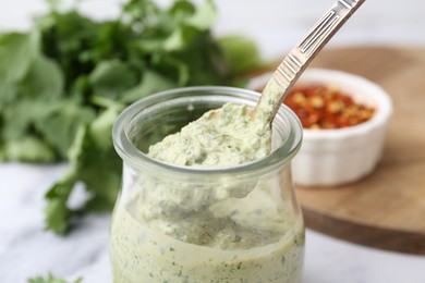 Photo of Taking tasty cilantro sauce with spoon from jar at light table, closeup