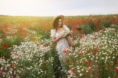 Photo of Smiling woman in hat with bag of chamomile flowers walking in wildflower meadow