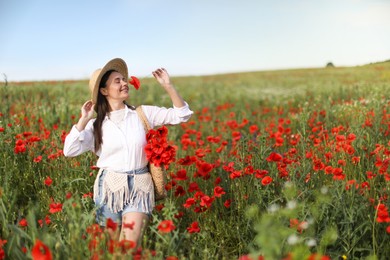 Photo of Smiling woman in hat with bag sniffing poppy flower in morning wildflower meadow