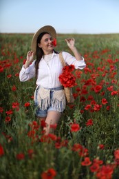 Photo of Smiling woman in hat with bag sniffing poppy flower in wildflower meadow