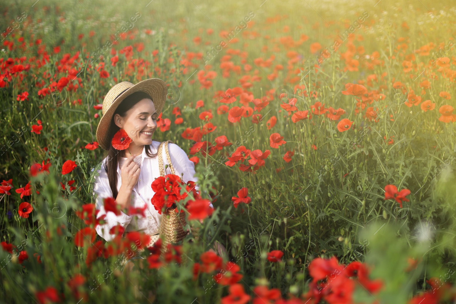 Photo of Smiling woman in hat with bag among red poppy flowers in morning wildflower meadow